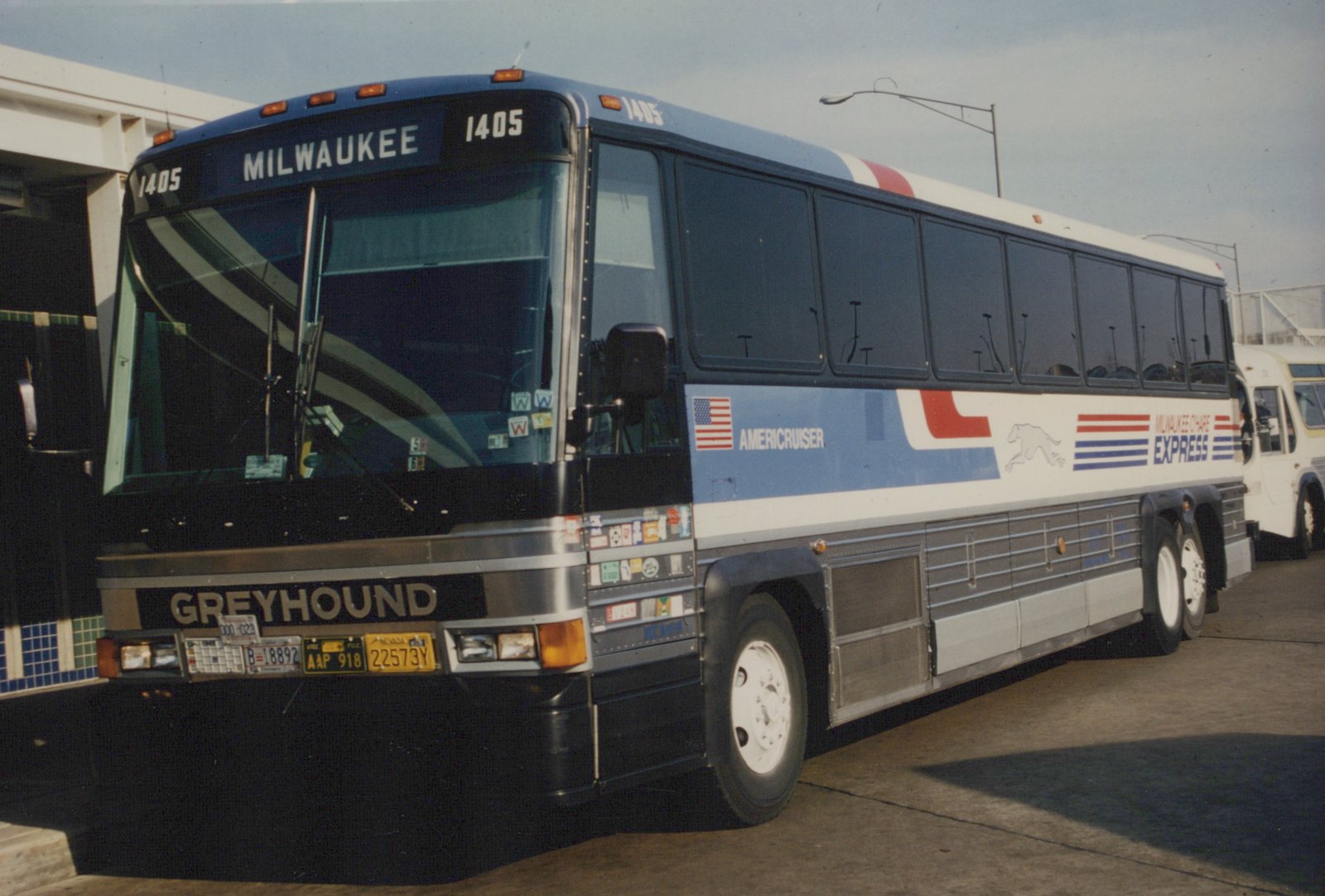 A Greyhound bus numbered 1405 with 'Milwaukee-O'Hare Express' lettering on its side, parked at a terminal, featuring a blue, white, and red livery with an American flag and a running greyhound logo.