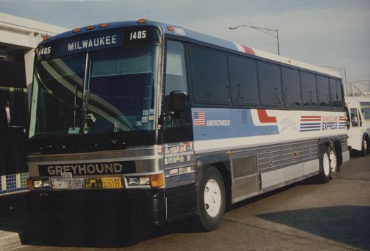 A Greyhound bus numbered 1405 with 'Milwaukee-O'Hare Express' lettering on its side, parked at a terminal, featuring a blue, white, and red livery with an American flag and a running greyhound logo.