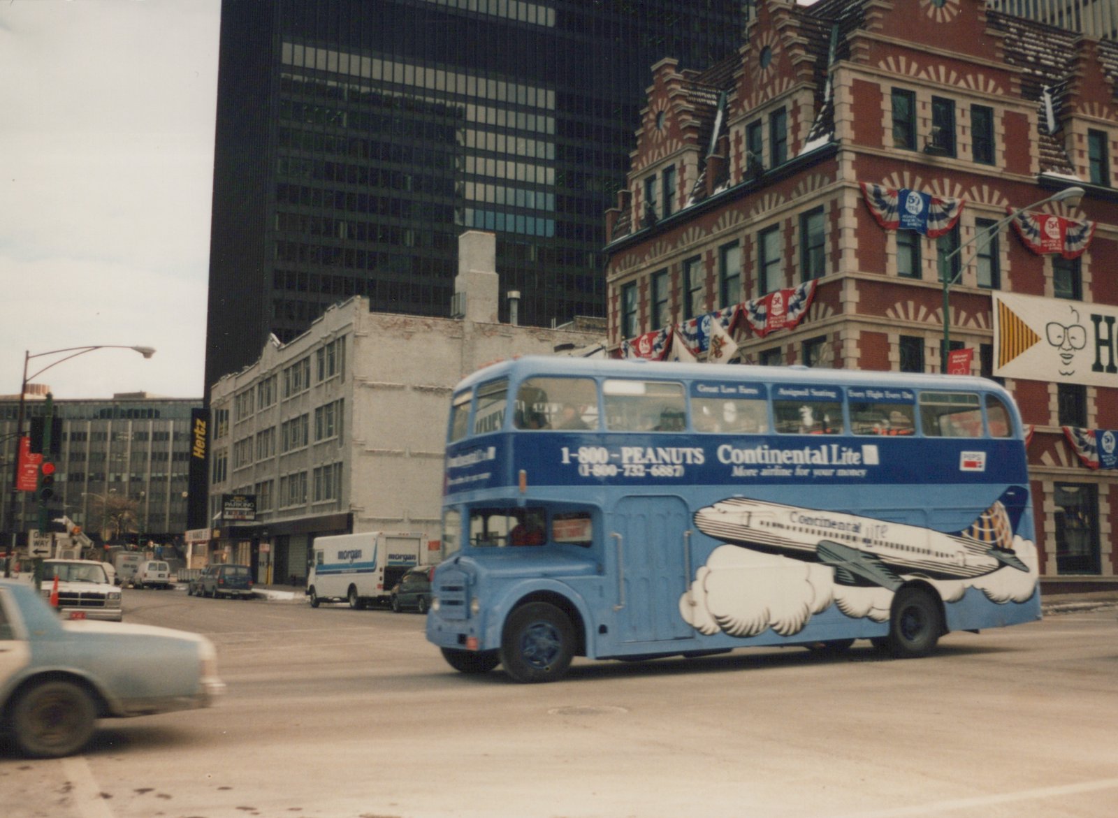 A light blue double-decker bus with a Continental Lite plane advertisement drives down a Chicago street, with a tall skyscraper and a red brick building in the background on an overcast day.