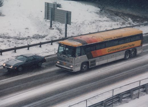 A silver and orange MCI motor coach with 'Continental Golden Charter' lettering drives on a snow-covered highway next to a dark car.