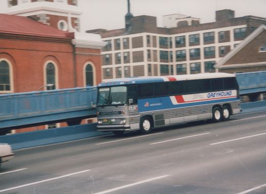 A blue, red and silver Greyhound bus is driving across a bridge under an overcast sky.