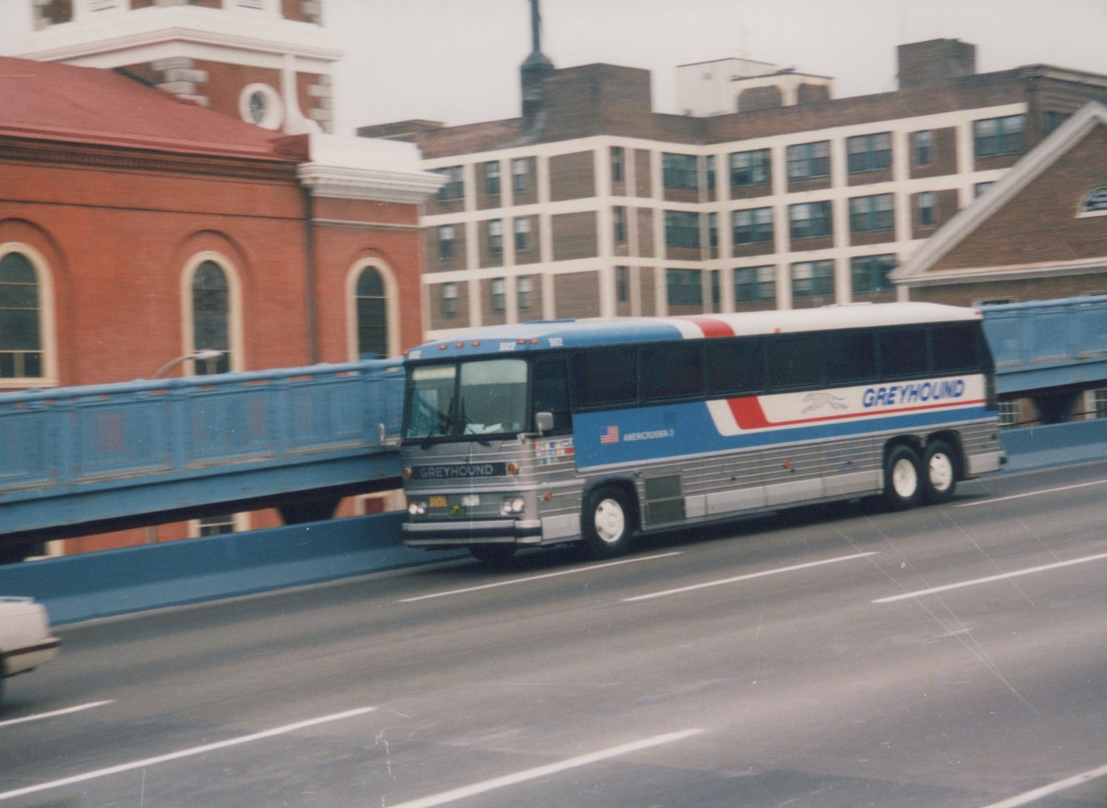 A blue, red and silver Greyhound bus is driving across a bridge under an overcast sky.