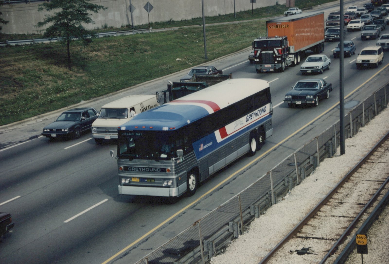 A long, blue, white, and red Greyhound bus numbered 1181 drives on a highway, viewed from above; cars and trucks are in adjacent lanes, and railroad tracks run along the right edge.