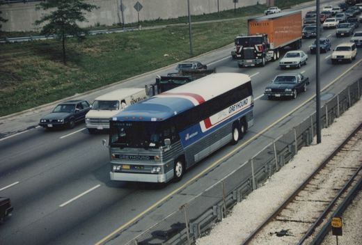 A long, blue, white, and red Greyhound bus numbered 1181 drives on a highway, viewed from above; cars and trucks are in adjacent lanes, and railroad tracks run along the right edge.