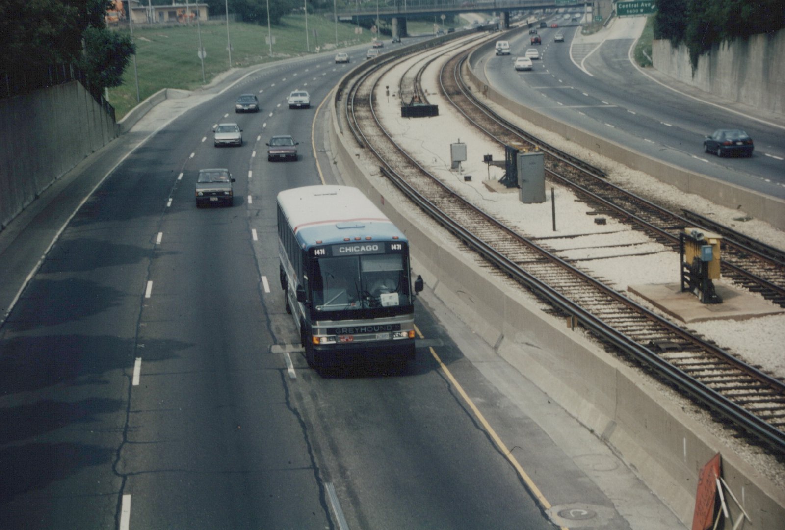 A Greyhound bus with number 1471 and destination 'Chicago' travels in the center median of a highway, with cars on either side and train tracks running parallel on its right.