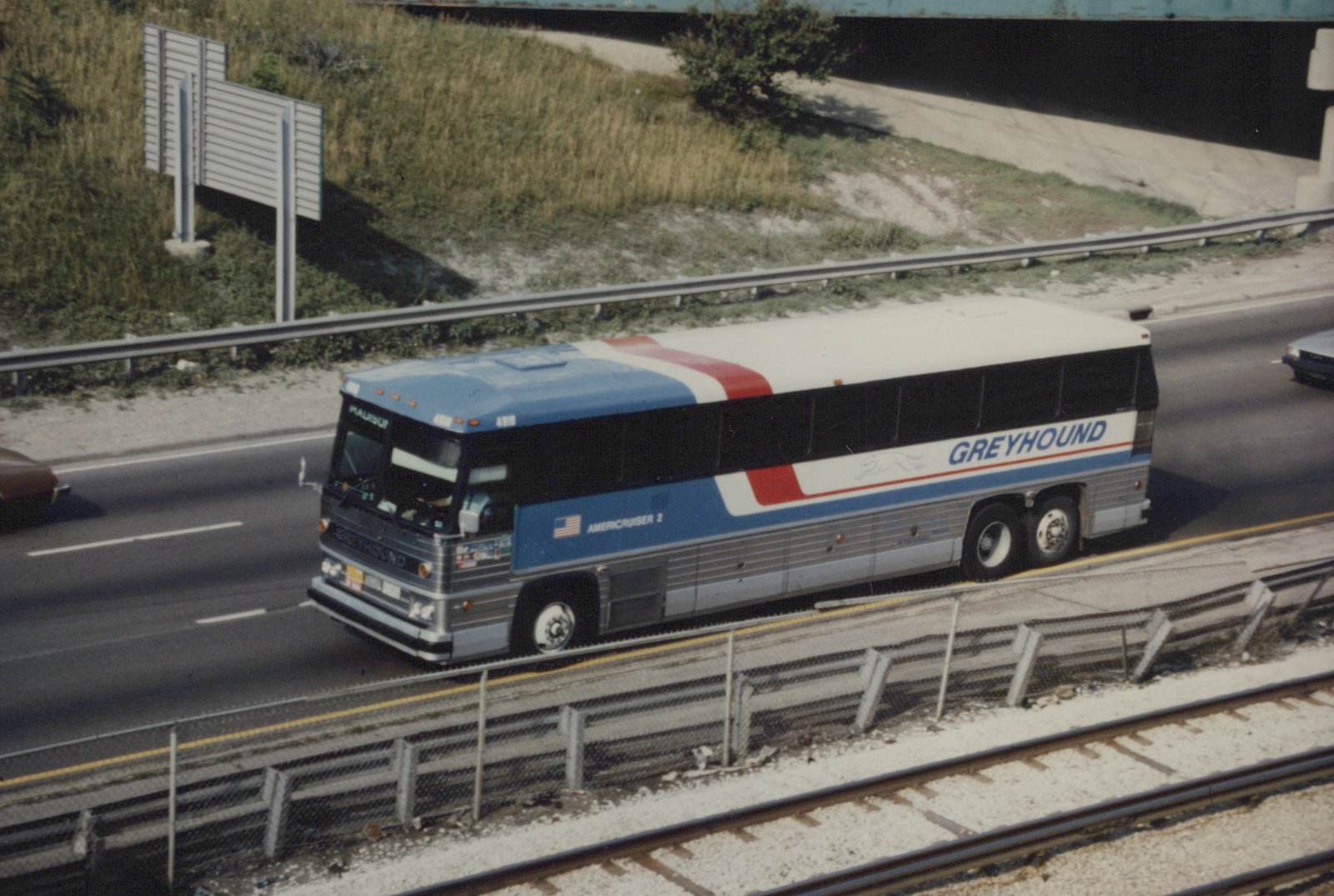 A large Greyhound bus with a blue, white, and red livery is driving on a multi-lane highway, viewed from above with railroad tracks visible below.
