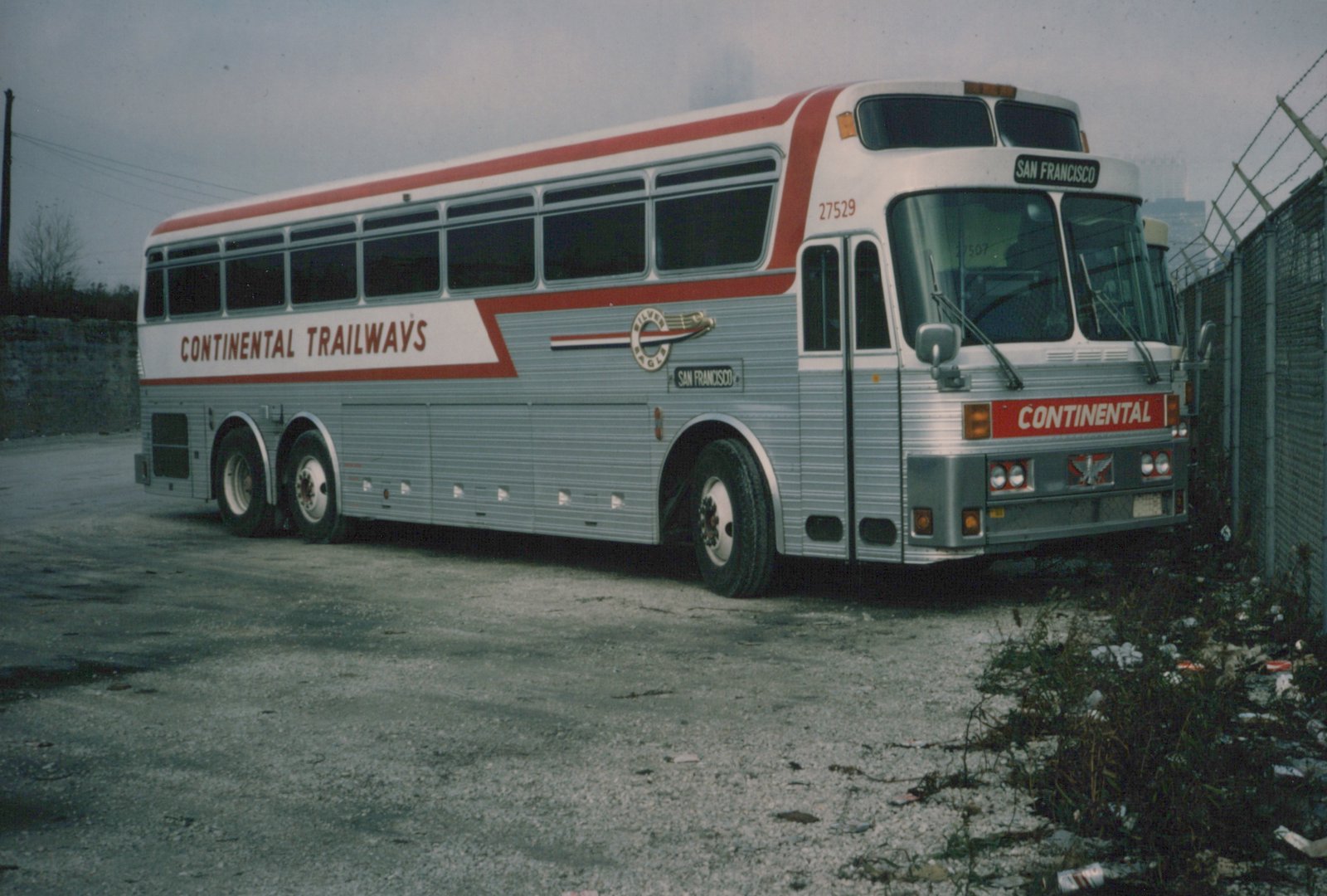 A silver and red Continental Trailways motor coach, number 27529, is parked in a paved lot next to a chain-link fence, with 'San Francisco' displayed on its front sign.