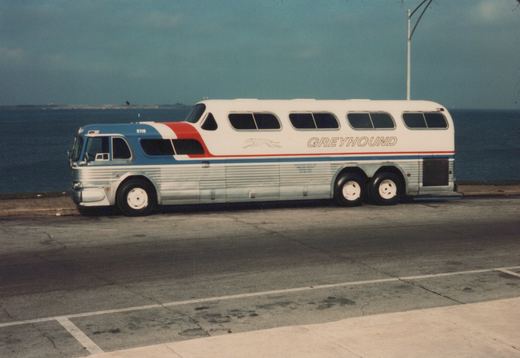 Side view of a white, blue, and red Greyhound Scenicruiser bus, fleet number 8718, parked on pavement next to water under a cloudy sky.