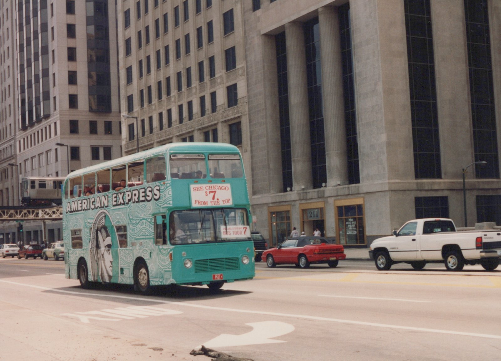 A turquoise American Express double-decker tour bus with the number 8511 is driving on a city street, with a red convertible and a white pickup truck nearby, and a silver 'L' train on an elevated track in the background.