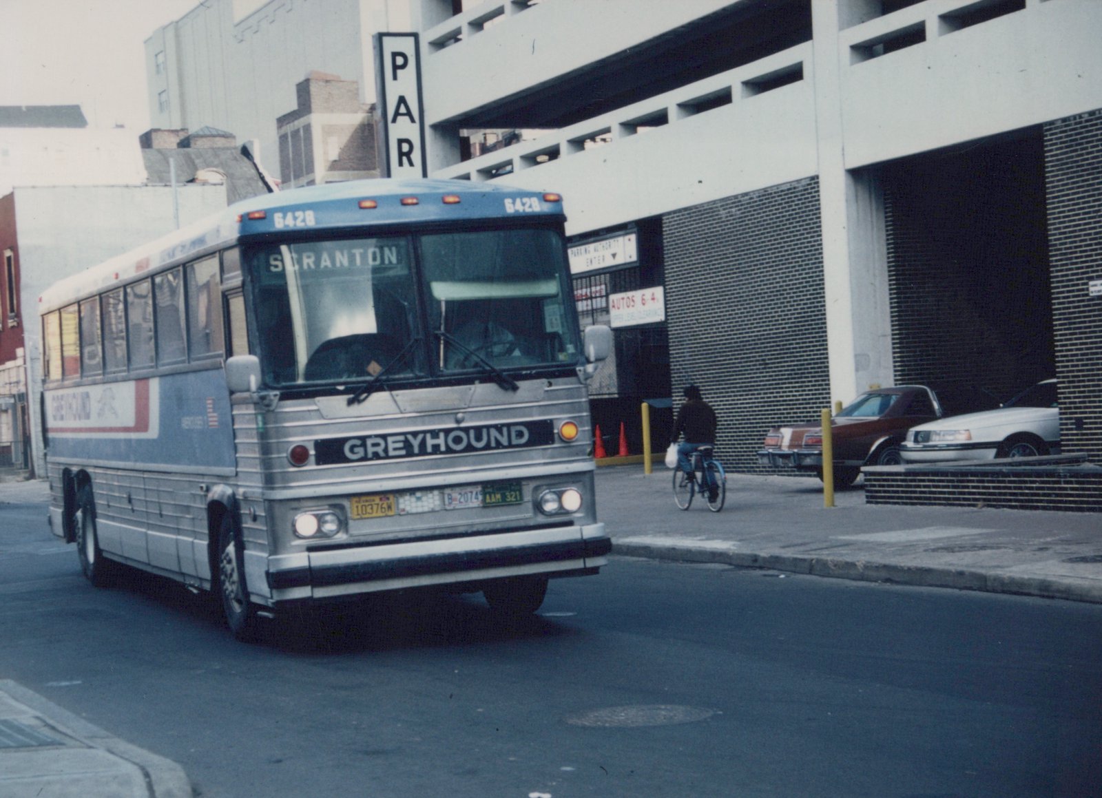 A silver and blue Greyhound motorcoach, fleet number 6428, with a 'Scranton' destination sign, is driving on a city street past a multi-story parking garage while a cyclist rides on the sidewalk.