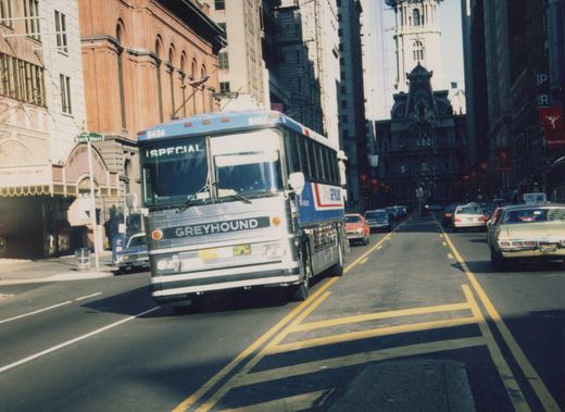 A white and blue Greyhound bus with the number 8434 is driving down a city street lined with cars and tall buildings, with a street sign reading 'Bach Place' visible on the left.