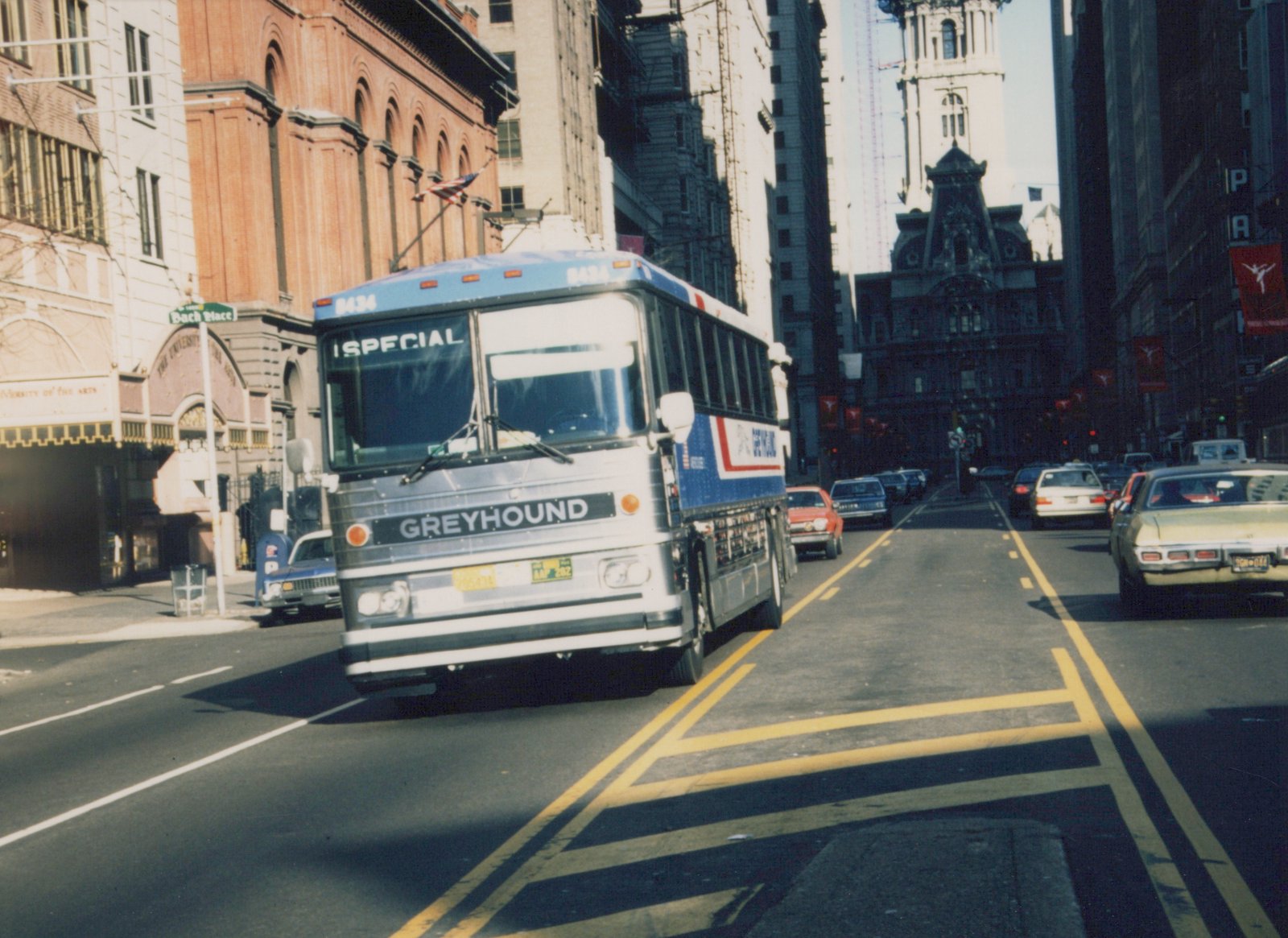 A white and blue Greyhound bus with the number 8434 is driving down a city street lined with cars and tall buildings, with a street sign reading 'Bach Place' visible on the left.