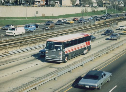 A long, gray and white motor coach with red stripes and the words 'Wisconsin-Michigan Trailways' on the side drives on a highway median next to railroad tracks, with 1970s and 80s cars in nearby lanes.