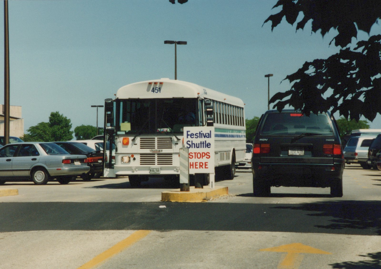 A white Pace bus numbered 451 is stopped at a 'Festival Shuttle STOPS HERE' sign in a parking lot on a sunny day, with a black SUV and other cars nearby.