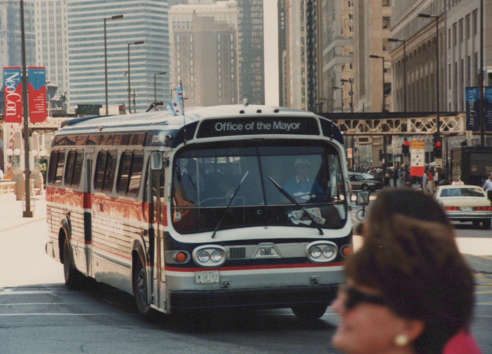 A white, red, and blue city bus with 'Office of the Mayor' on the front sign drives down a city street with a pedestrian in the foreground and tall buildings and an elevated train structure in the background.