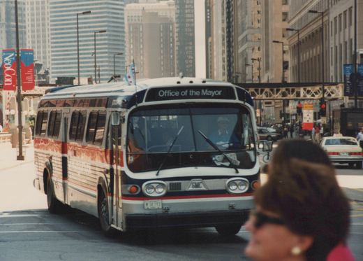 A white, red, and blue city bus with 'Office of the Mayor' on the front sign drives down a city street with a pedestrian in the foreground and tall buildings and an elevated train structure in the background.