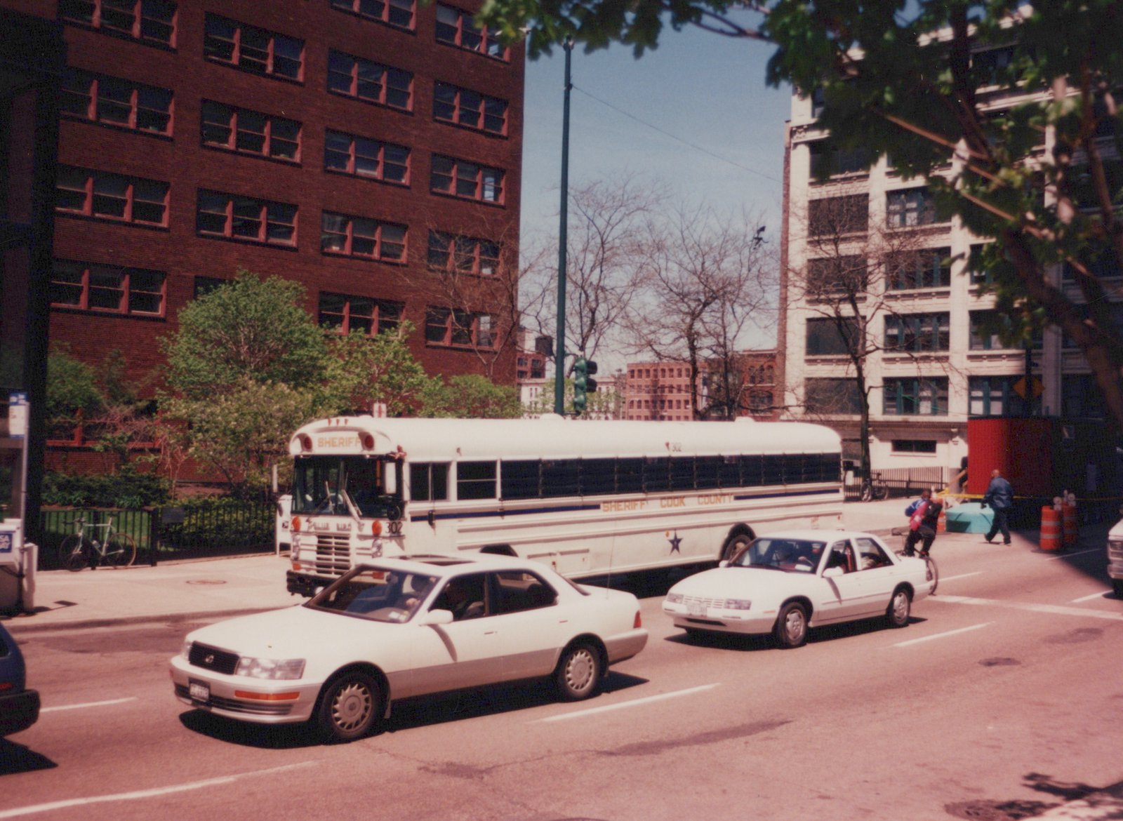 A white Cook County Sheriff's bus numbered 302, a Pullman 818A model, is parked or moving slowly on a city street with two white sedans in the foreground and pedestrians nearby.