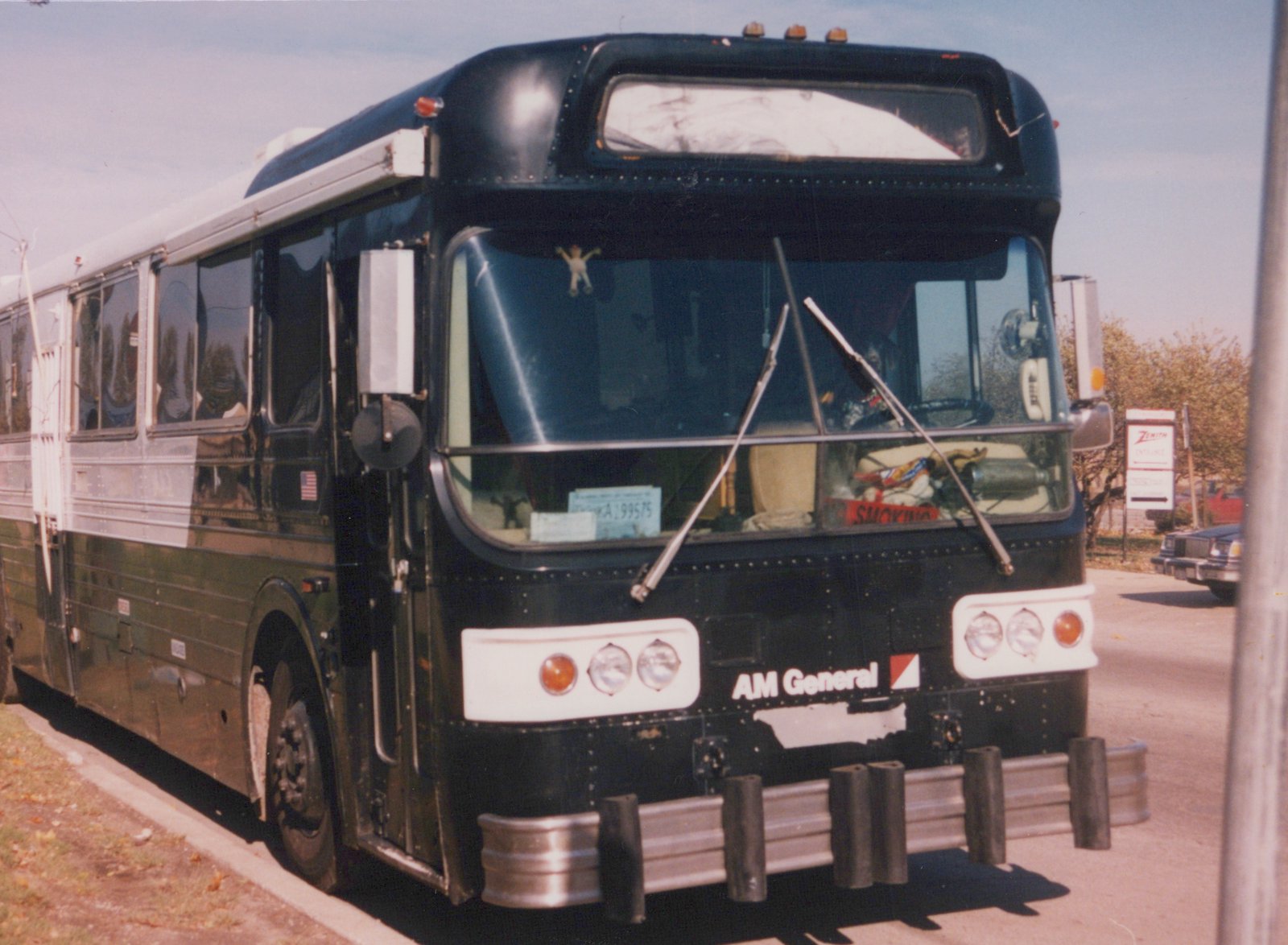 A black and silver AM General motor bus is parked on a street, with an autumnal background and a clear sky.