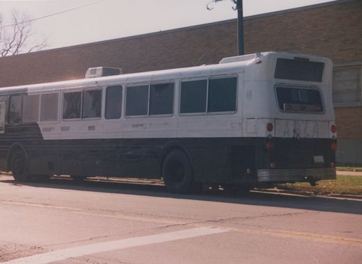 A white and black motor bus with fleet number 32005 on its rear license plate, parked on the side of a street next to a brick building with bare trees in the background.