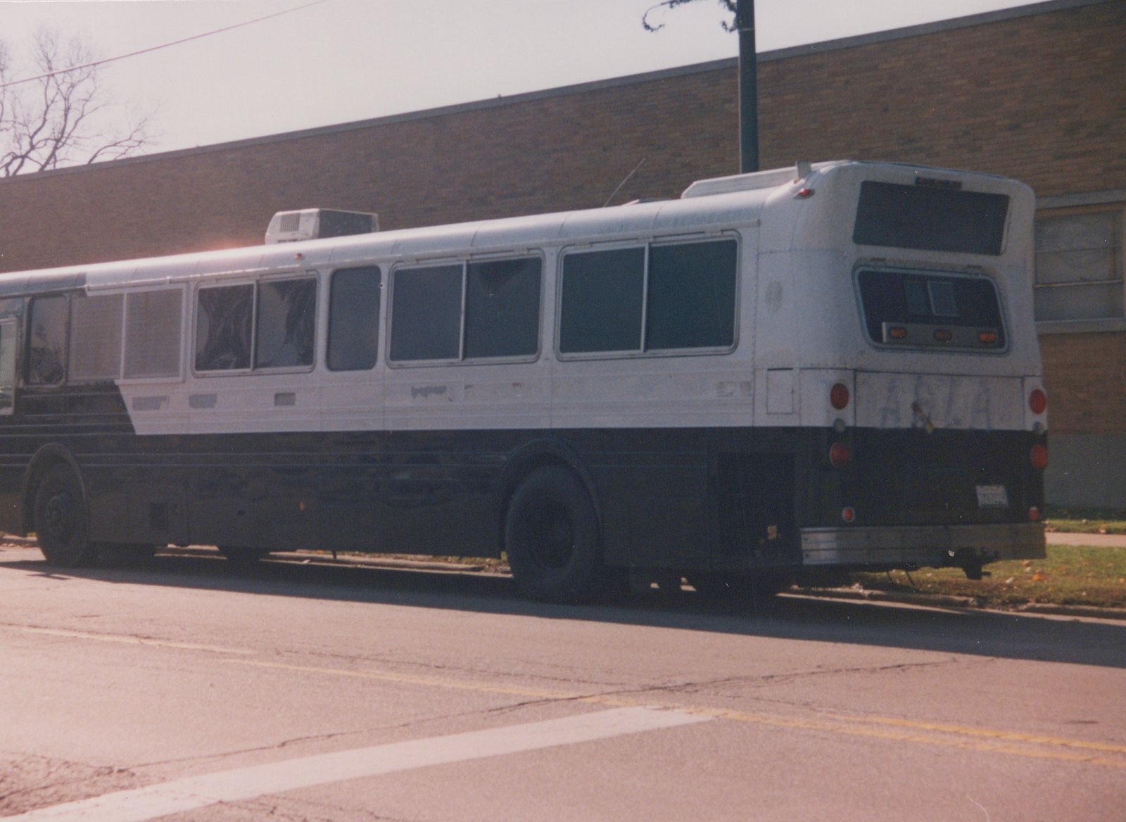 A white and black motor bus with fleet number 32005 on its rear license plate, parked on the side of a street next to a brick building with bare trees in the background.