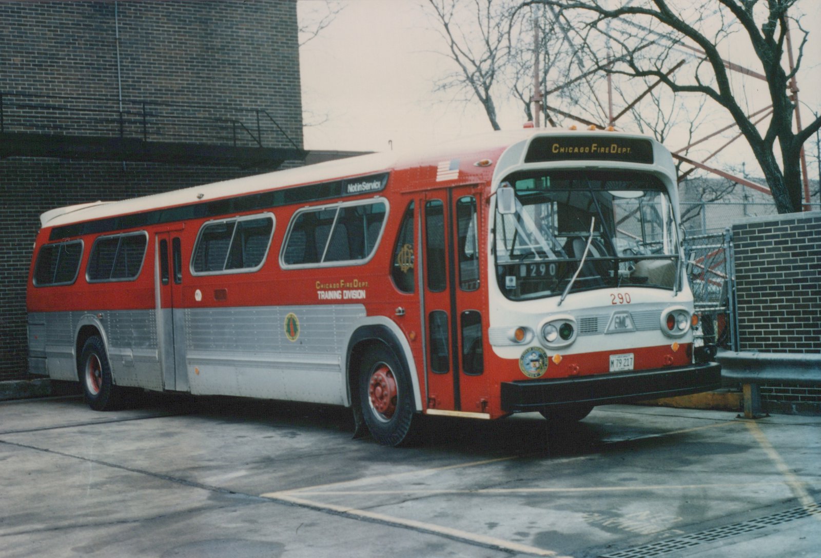 A red and white GMC bus with 'Chicago Fire Dept. Training Division' lettering and fleet number 290, parked in a lot next to a brick building.