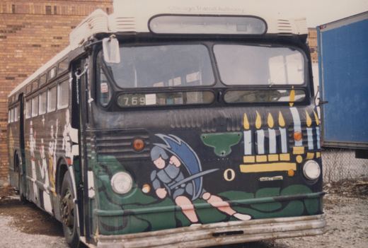 A vintage Chicago Transit Authority bus, number 769, painted with a holiday-themed design of a cartoon knight and a menorah, is parked on a gravel lot next to a brick building.