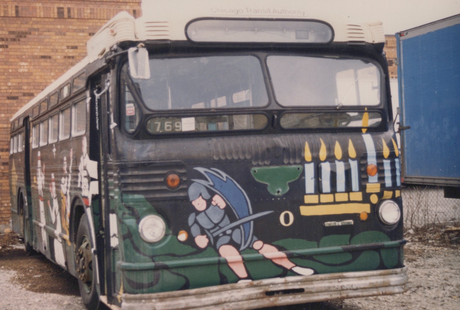 A vintage Chicago Transit Authority bus, number 769, painted with a holiday-themed design of a cartoon knight and a menorah, is parked on a gravel lot next to a brick building.
