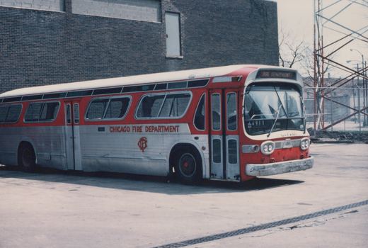 A red and silver Chicago Fire Department bus, number 911, parked on a paved lot with a brick building in the background.