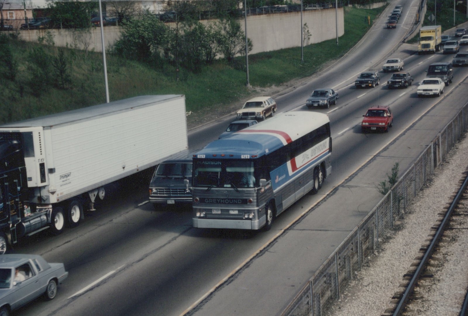 A Greyhound bus with the number 6253 and destination 'MADISON' is shown driving on a multi-lane highway alongside other vehicles, with a freight train track visible on the right and a concrete retaining wall in the background.