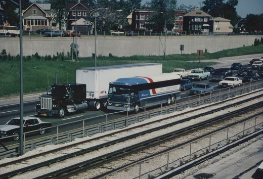 A Greyhound bus with number 7824 drives on a highway next to train tracks, surrounded by other vehicles and residential houses in the background.
