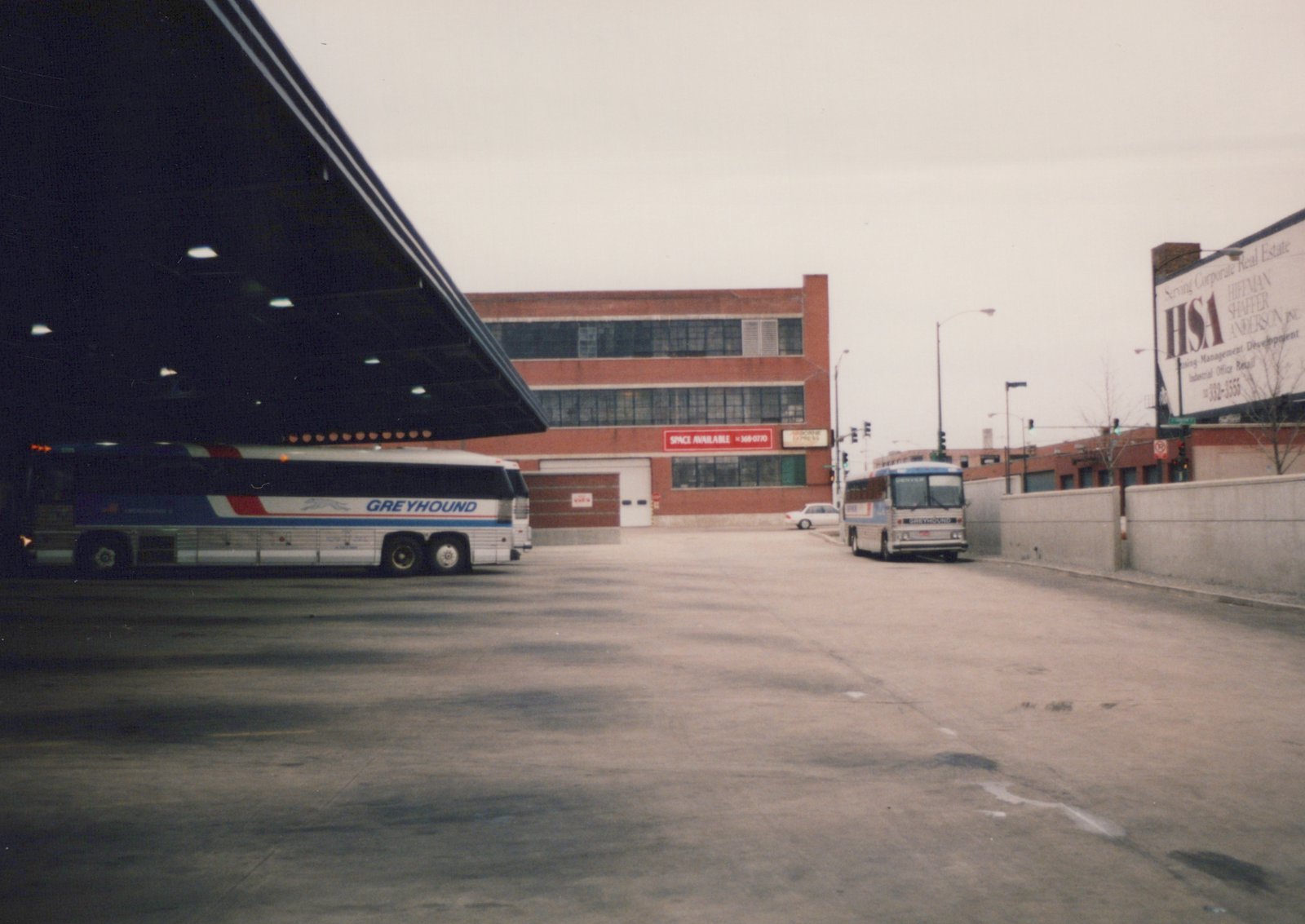 Two Greyhound buses, one parked under a shelter and another on the street, are seen in a drab, overcast urban scene with a brick building and a large billboard in the background.
