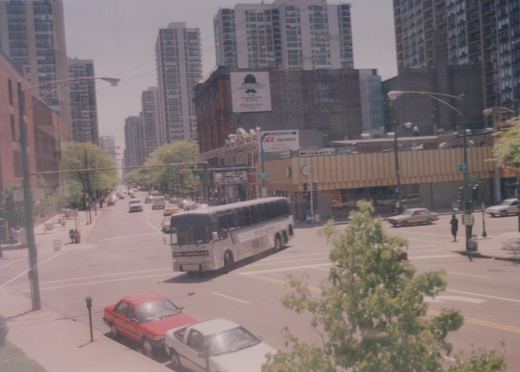 A white Mid-America motor bus drives through a city street intersection with tall buildings in the background and cars parked along the curb.