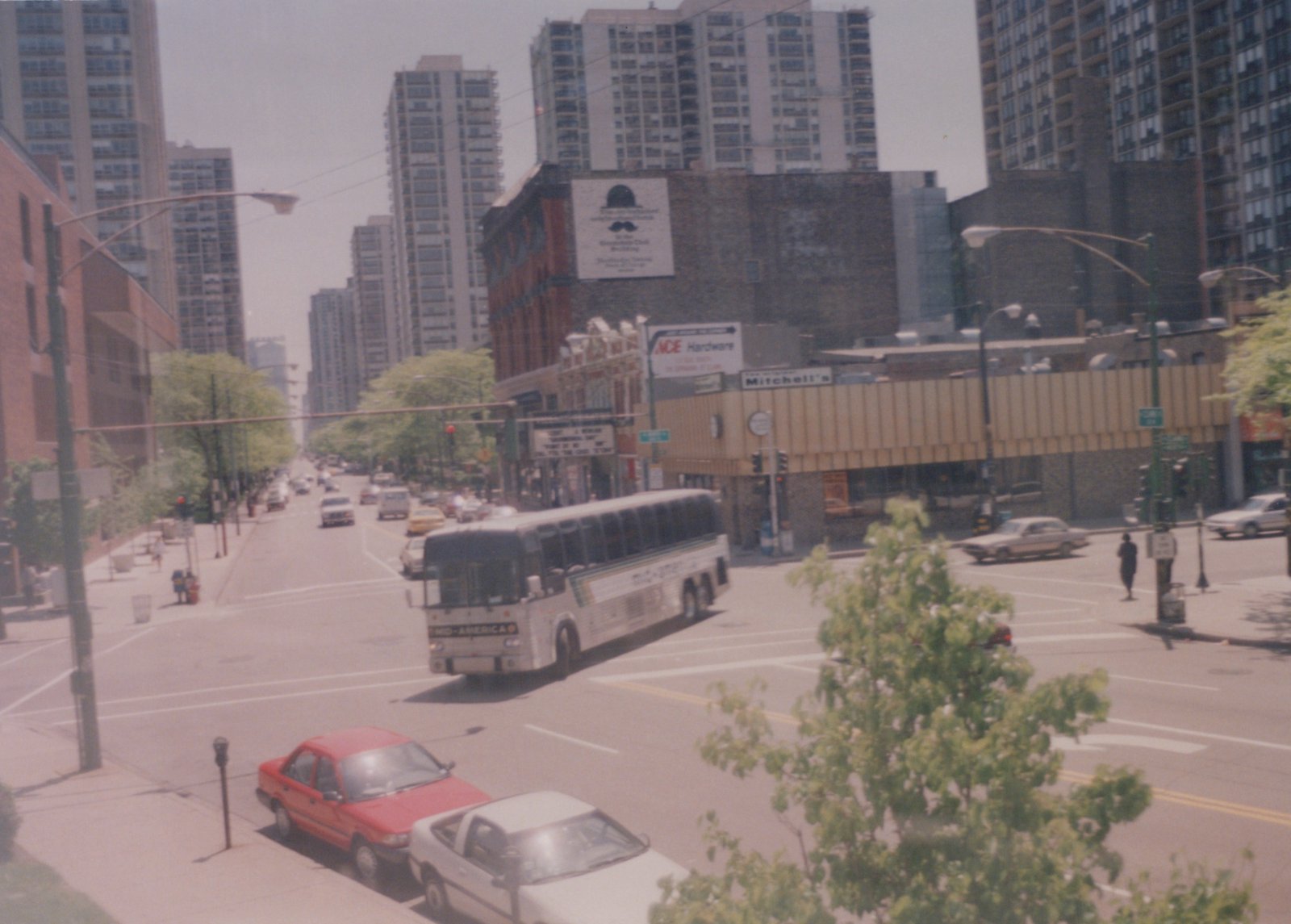 A white Mid-America motor bus drives through a city street intersection with tall buildings in the background and cars parked along the curb.
