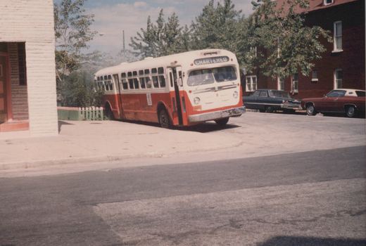 A red and white Flxible bus, number 8, labeled 'CHARTERED', is parked on a street with two vintage cars in the background under a clear sky.