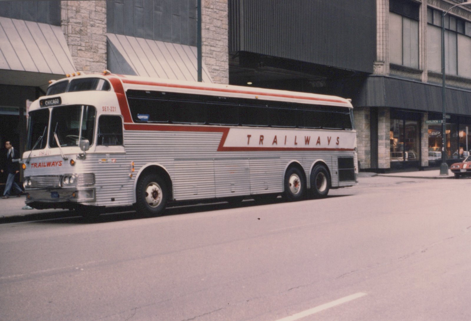A silver and red Trailways motor bus, SET-221, parked on a city street with a pedestrian walking by and a building in the background.