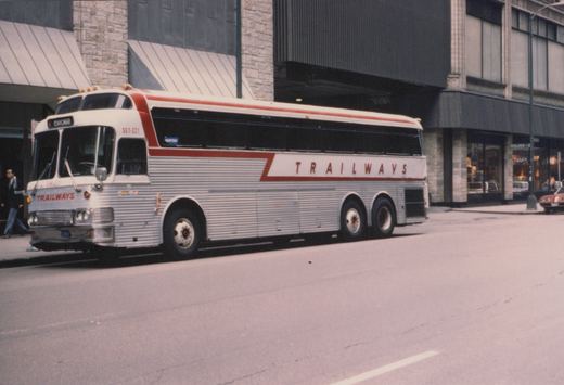A silver and red Trailways motor bus, SET-221, parked on a city street with a pedestrian walking by and a building in the background.