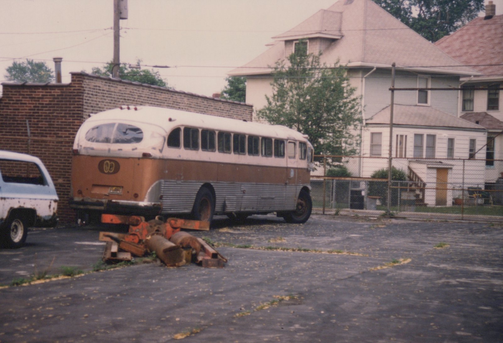 A vintage orange and white bus with a 'CC' emblem on the back is parked in a lot next to a rusted white pickup truck, with houses and a fence in the background.