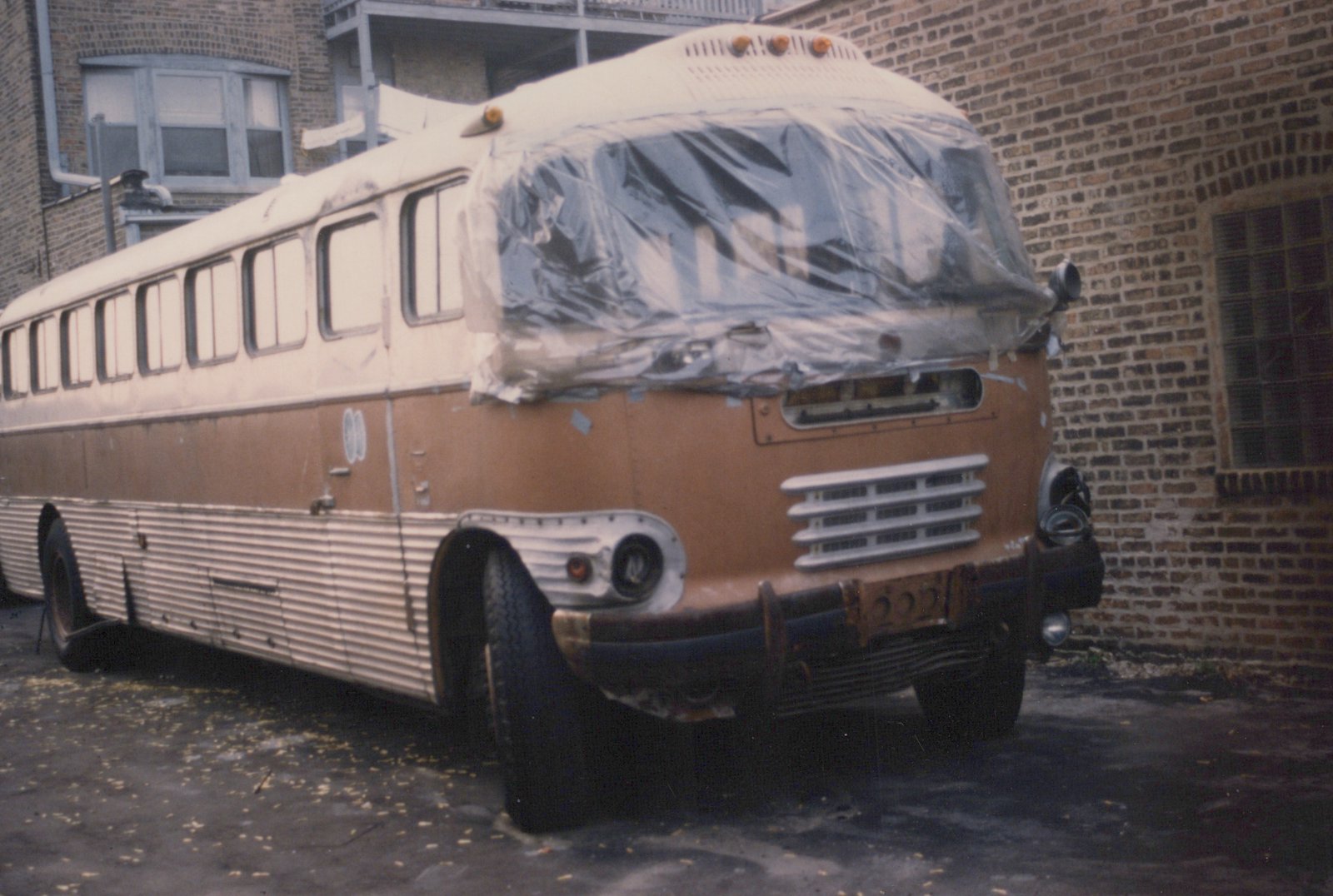 A vintage brown and cream bus with a plastic-covered windshield is parked on an asphalt surface next to a brick building.
