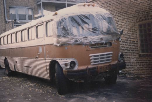 A vintage brown and cream bus with a plastic-covered windshield is parked on an asphalt surface next to a brick building.