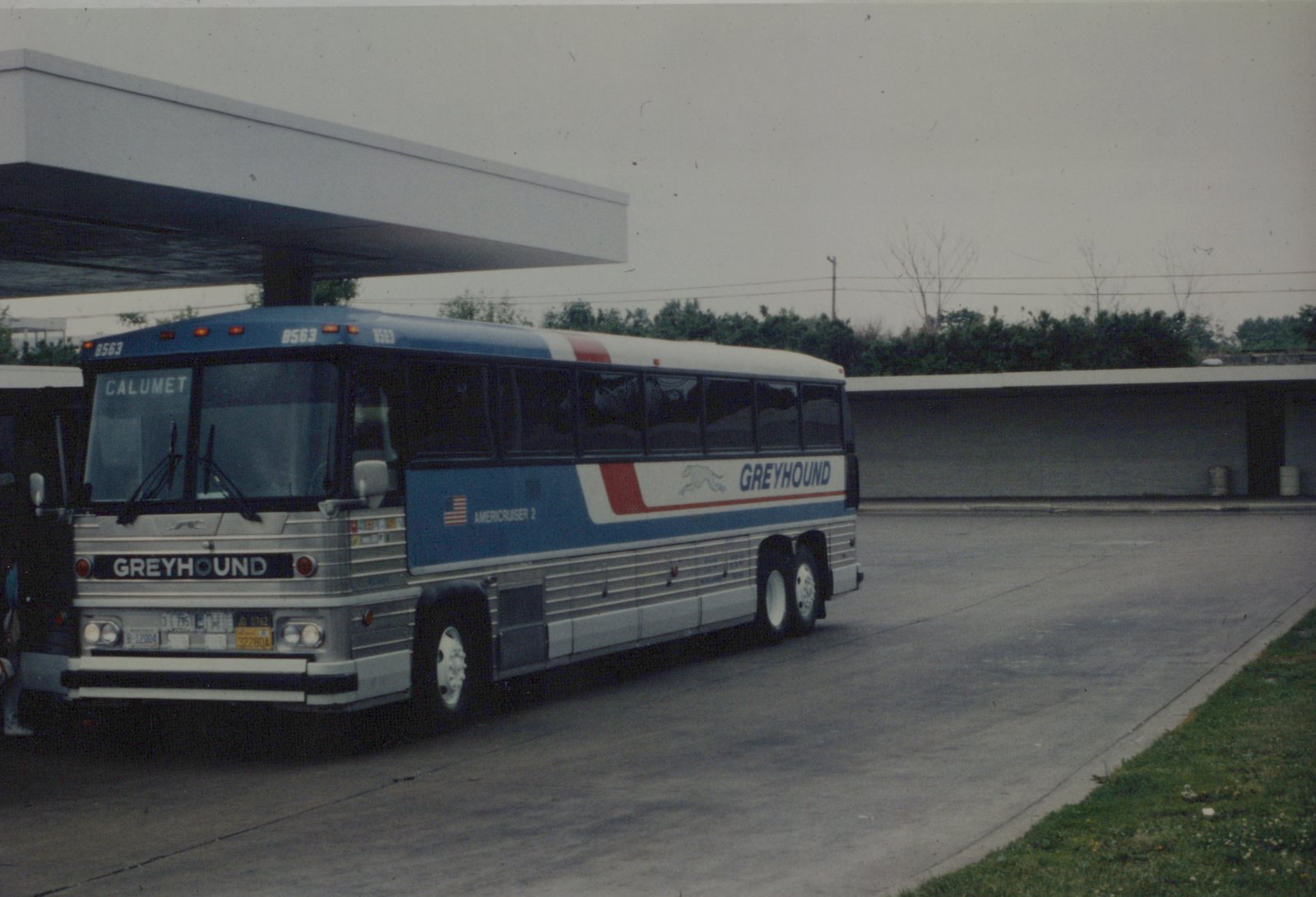A blue, white, and red Greyhound motor bus, number 8563, is parked under a gas station canopy, with a destination sign reading 'CALUMET'.