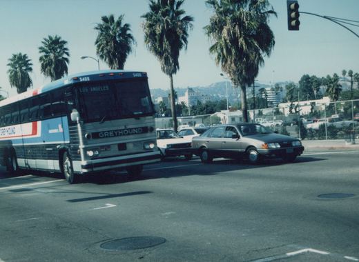 A large Greyhound coach, number 5408, with a destination sign reading 'LOS ANGELES' is stopped at a street intersection, surrounded by cars and tall palm trees under a clear blue sky.