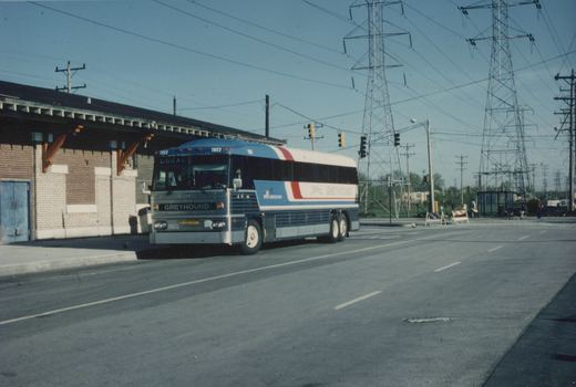 A blue, white, and red Greyhound bus, number 7853, is stopped on a street corner near a brick building, with large power lines and towers in the background.