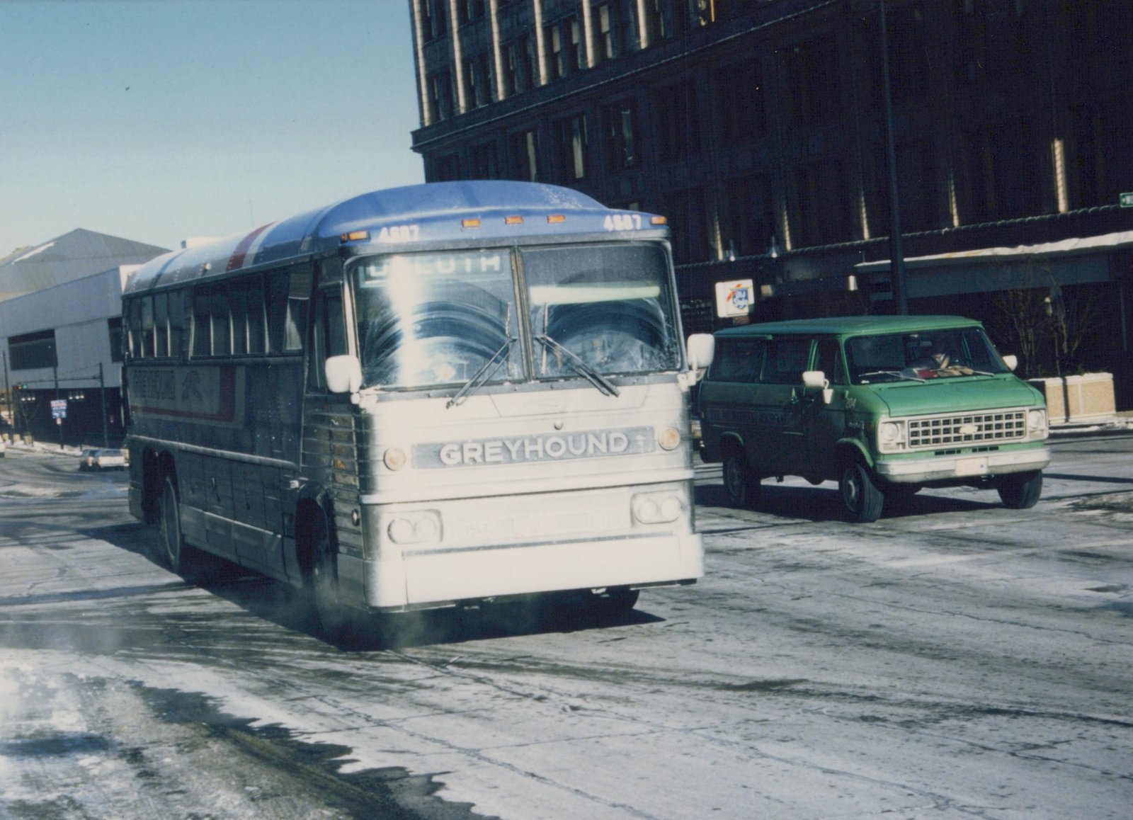 A white and blue Greyhound bus with the number 4687 on its roof travels down a snow-dusted city street, with a green van beside it and tall buildings in the background.