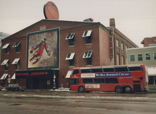 A red double-decker bus is parked on a wet, snowy street in front of a brick building with a large Michael Jordan mural and a sign for Michael Jordan's Restaurant.