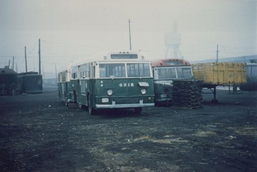 A green and cream Chicago Transit Authority bus numbered 4318 is parked in a gravel yard. Behind it is a red and white bus, a yellow freight car on a flatcar, and a dark railcar to the left. Overhead wires and a water tower are visible in the background under an overcast sky.
