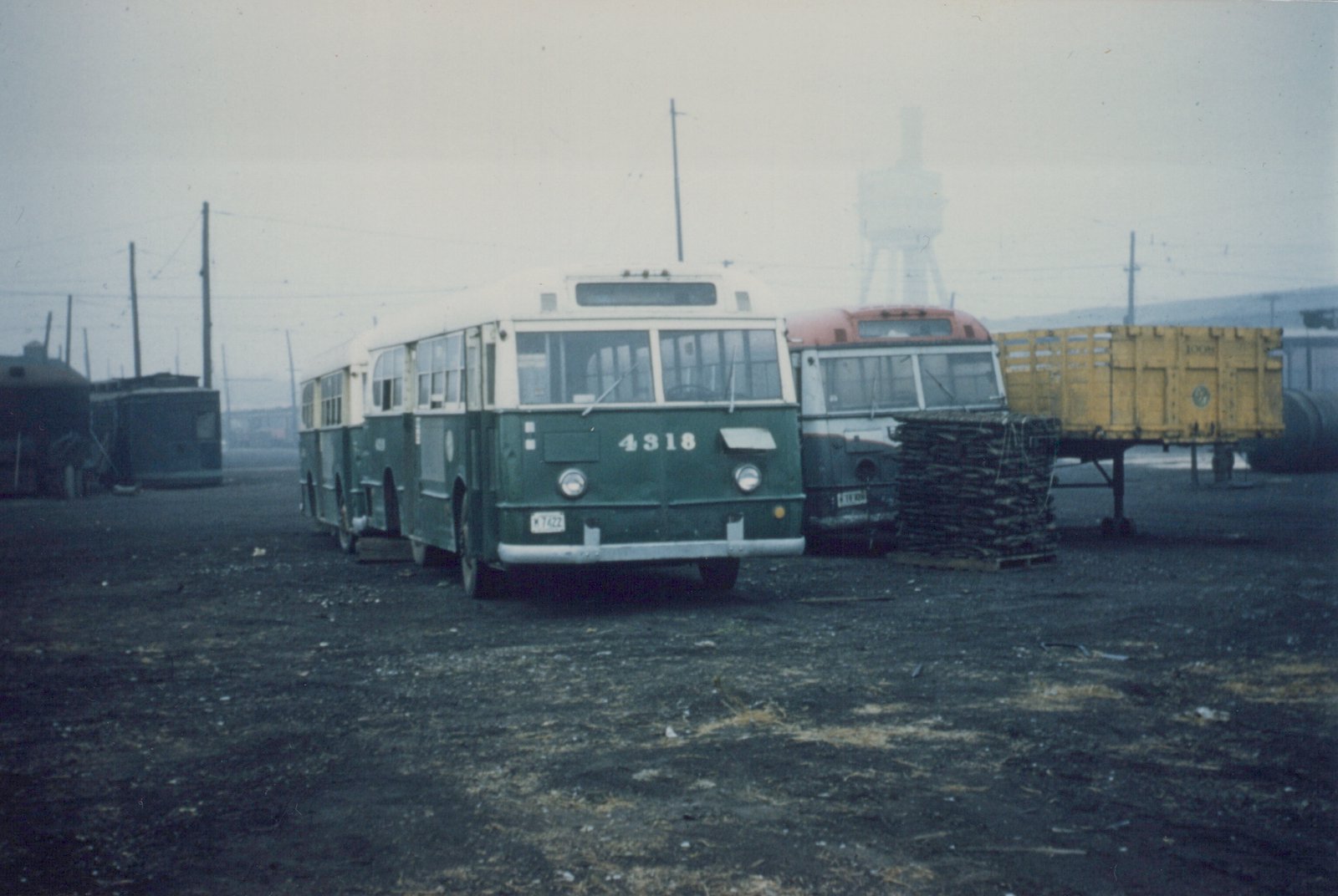A green and cream Chicago Transit Authority bus numbered 4318 is parked in a gravel yard. Behind it is a red and white bus, a yellow freight car on a flatcar, and a dark railcar to the left. Overhead wires and a water tower are visible in the background under an overcast sky.