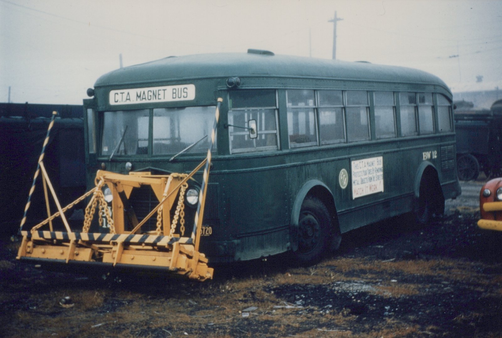 A dark green CTA bus with a yellow magnet sweeper attachment on its front, parked in a yard with a freight car visible behind it.