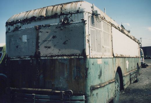 Rear three-quarter view of a decayed white and teal bus with rusted body panels and a damaged roof, parked in a yard.