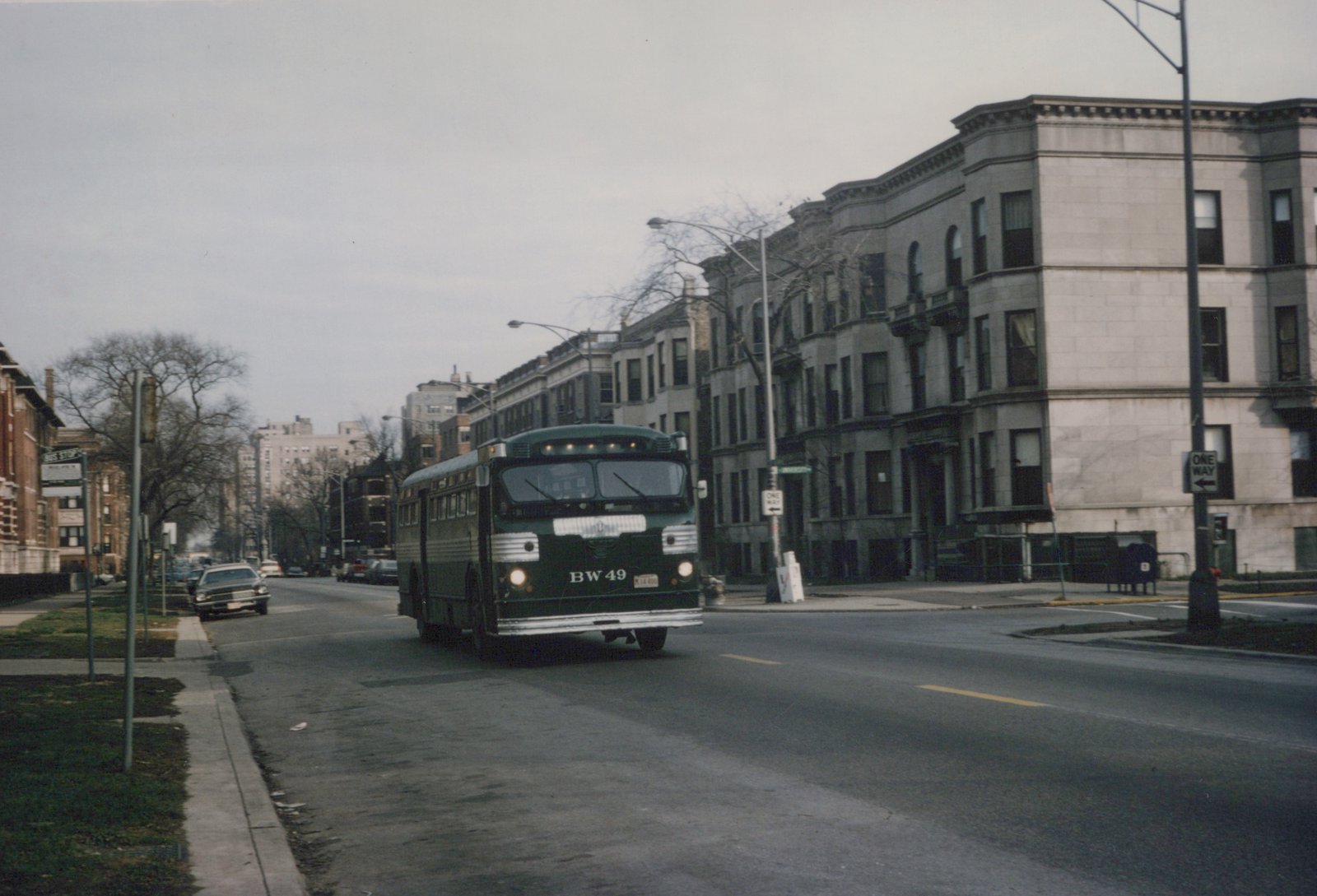 A dark green motor bus with fleet number BW 49 drives down a street lined with buildings and bare trees under an overcast sky.