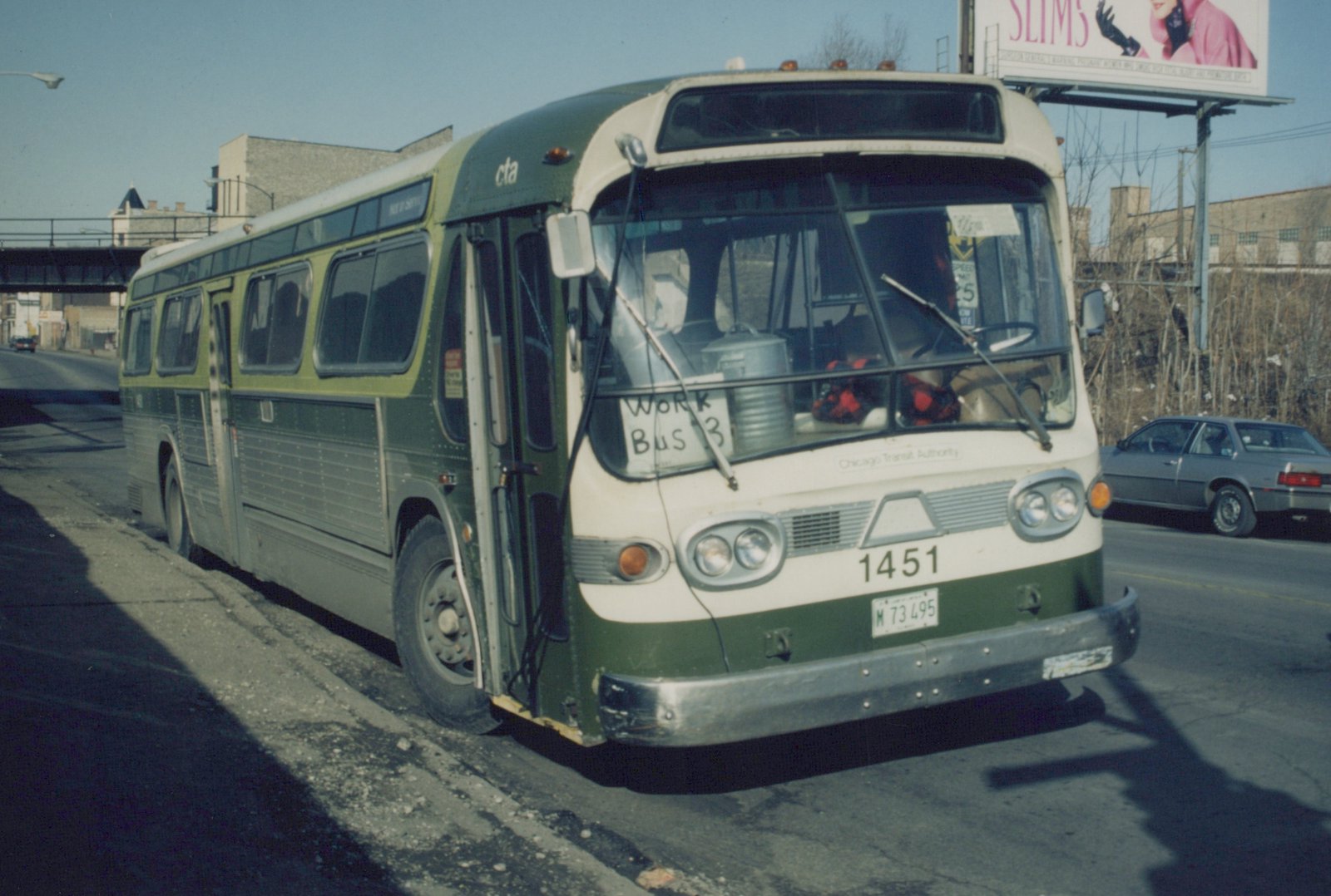 A green and cream CTA motor bus, number 1451, is parked on a city street in winter, with a handwritten 'Work Bus 3' sign on its windshield and a silver car in the background.